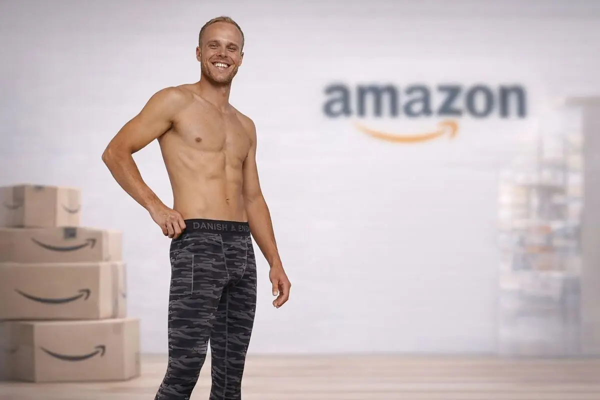 Hombre sonriente sin camiseta posando con mallas deportivas de camuflaje frente a cajas de envío y un gran logo de Amazon desenfocado al fondo