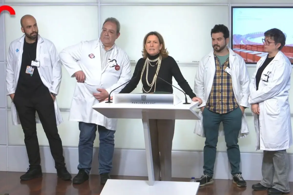 Grup de professionals sanitaris amb bata blanca acompanyant una dona que parla en un faristol durant una roda de premsa en un edifici institucional