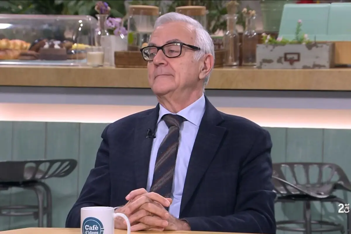 Hombre mayor con gafas y traje oscuro sentado en una cafetería de estilo moderno durante una entrevista televisiva