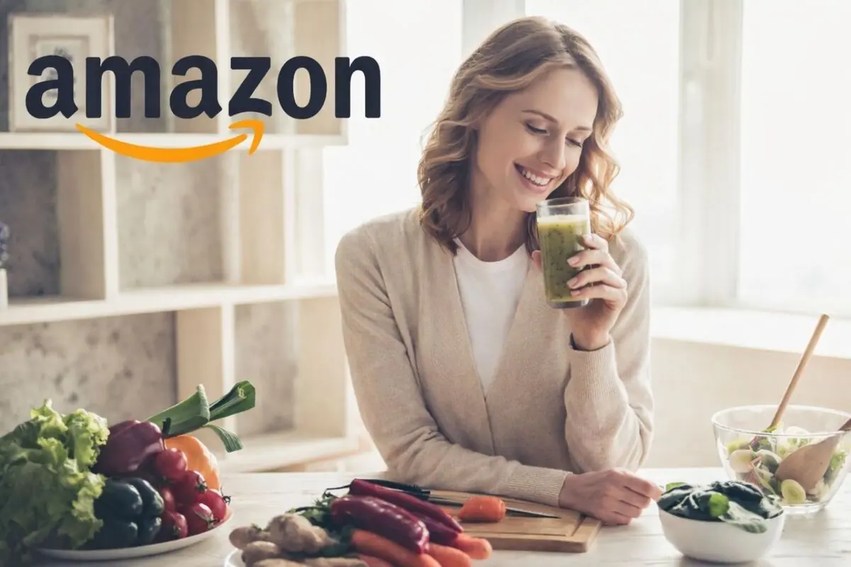 Mujer sonriendo mientras bebe un batido verde en una cocina con verduras frescas y el logo de Amazon en la parte superior izquierda