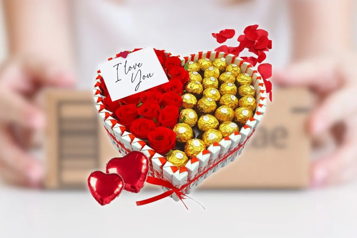 Montaje con corazón con rosas rojas y chocolates dorados Ferrero Rocher decorado con globos de corazón rojos y una tarjeta que dice I love you, teniendo de fondo una caja desenfocada