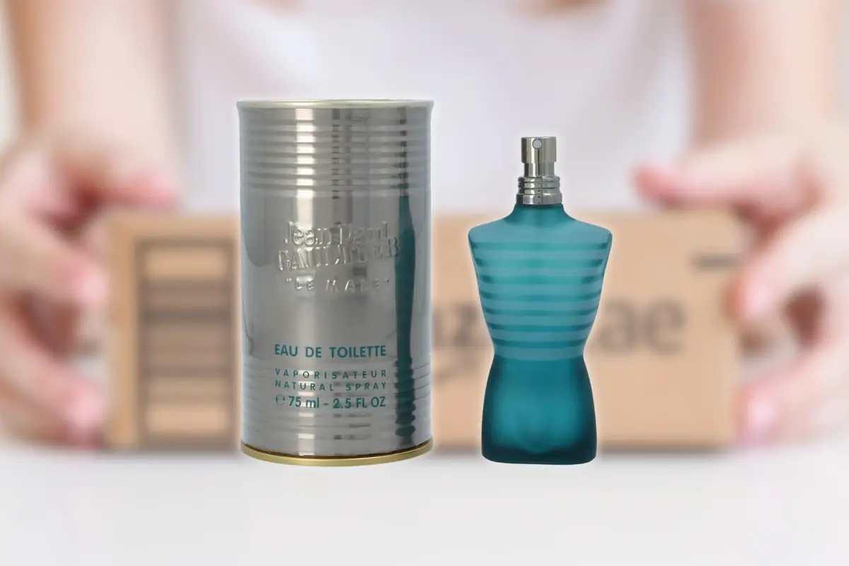 Montaje con perfume Jean Paul Gaultier Le Male con frasco azul en forma de torso masculino junto a su lata metálica plateada sobre fondo desenfocado de unas manos sujetando una caja