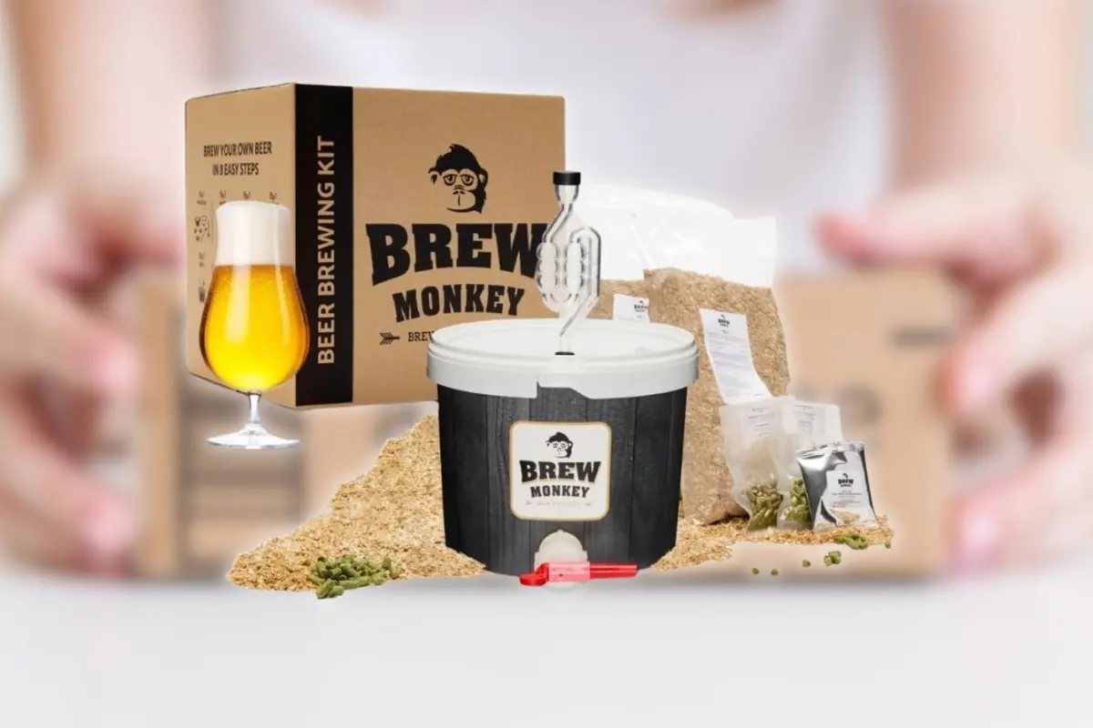 Montaje con Kit casero para hacer cerveza Brew Monkey con cubo fermentador, caja de embalaje, bolsas de malta y lúpulo y vaso de cerveza rubia servido junto a un fondo de unas manos desenfocadas sujetando caja