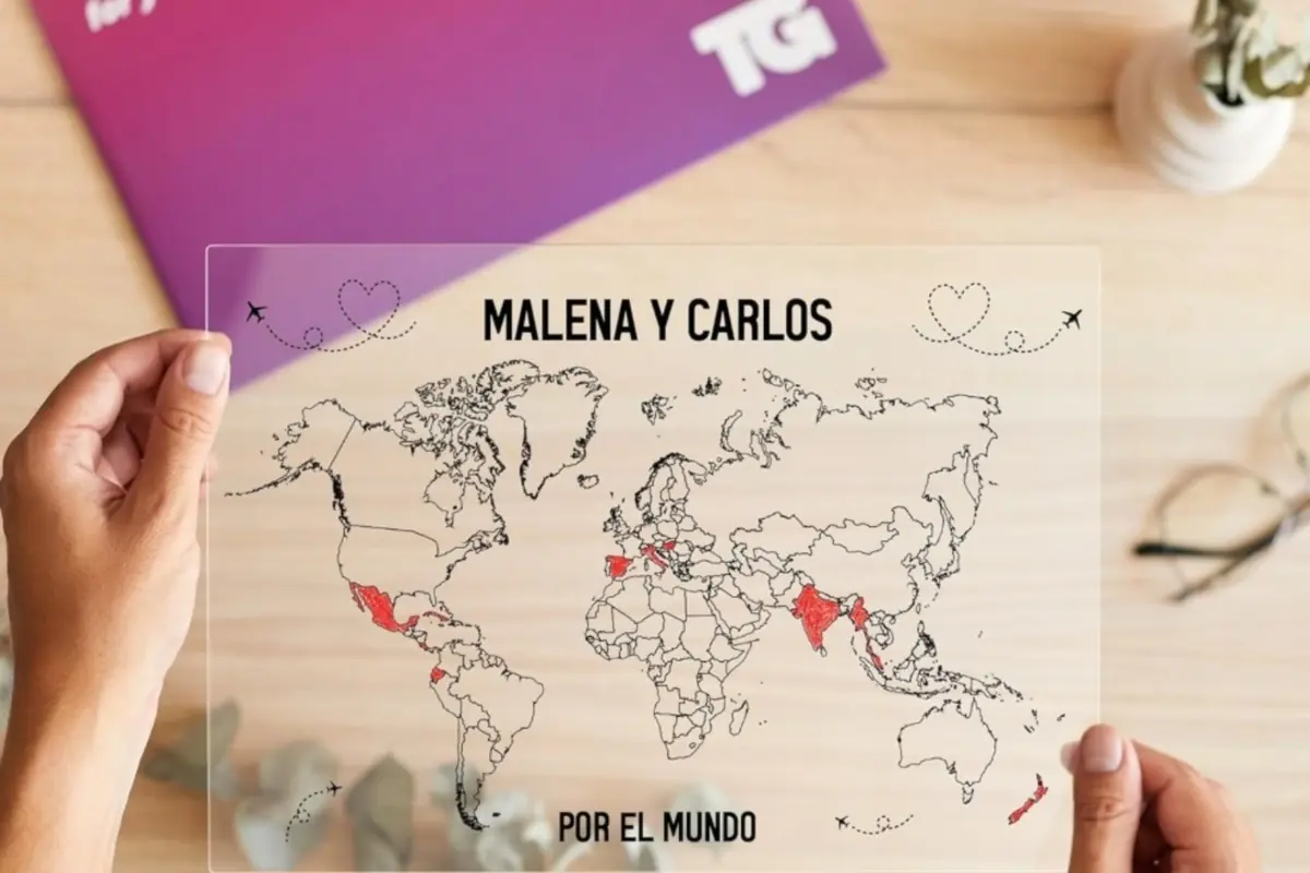 Manos sosteniendo una lámina transparente con mapa del mundo y países en rojo personalizada con el texto Malena y Carlos por el mundo sobre un escritorio de madera Manos sosteniendo una lámina transparente con mapa del mundo y países en rojo personalizada con el texto Malena y Carlos por el mundo sobre un escritorio de madera