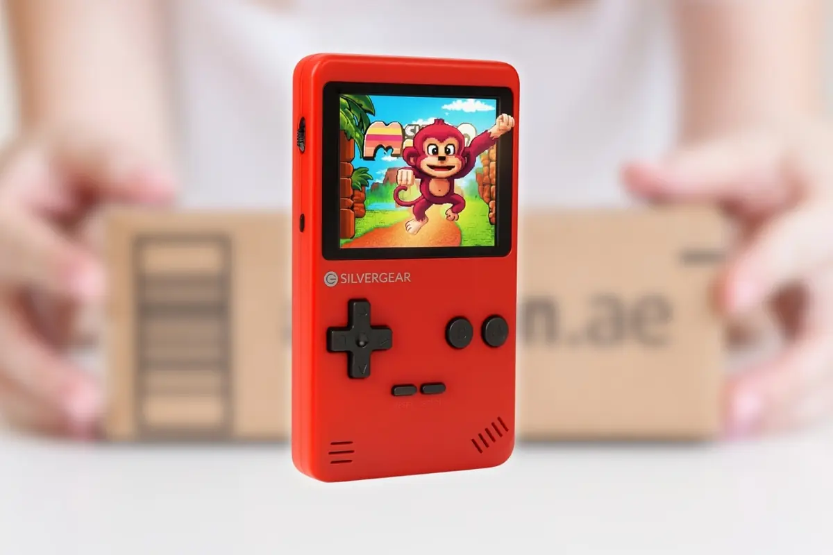 Montaje con consola portátil retro roja Silvergear con pantalla encendida mostrando un videojuego de mono saltando y fondo de paquete de envío desenfocado
