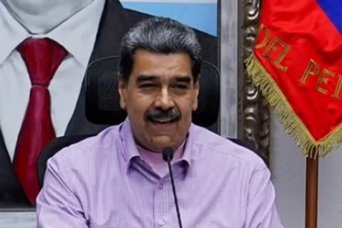 Pla mitjà de Nicolás Maduro amb camisa lila parlant davant d’un micròfon amb una bandera vermella al seu costat.