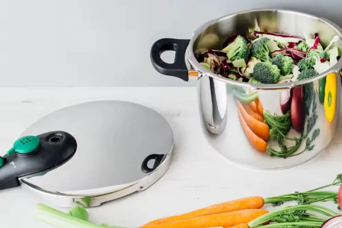 Olla a presión de acero inoxidable abierta con verduras frescas en su interior y más vegetales sobre la mesa