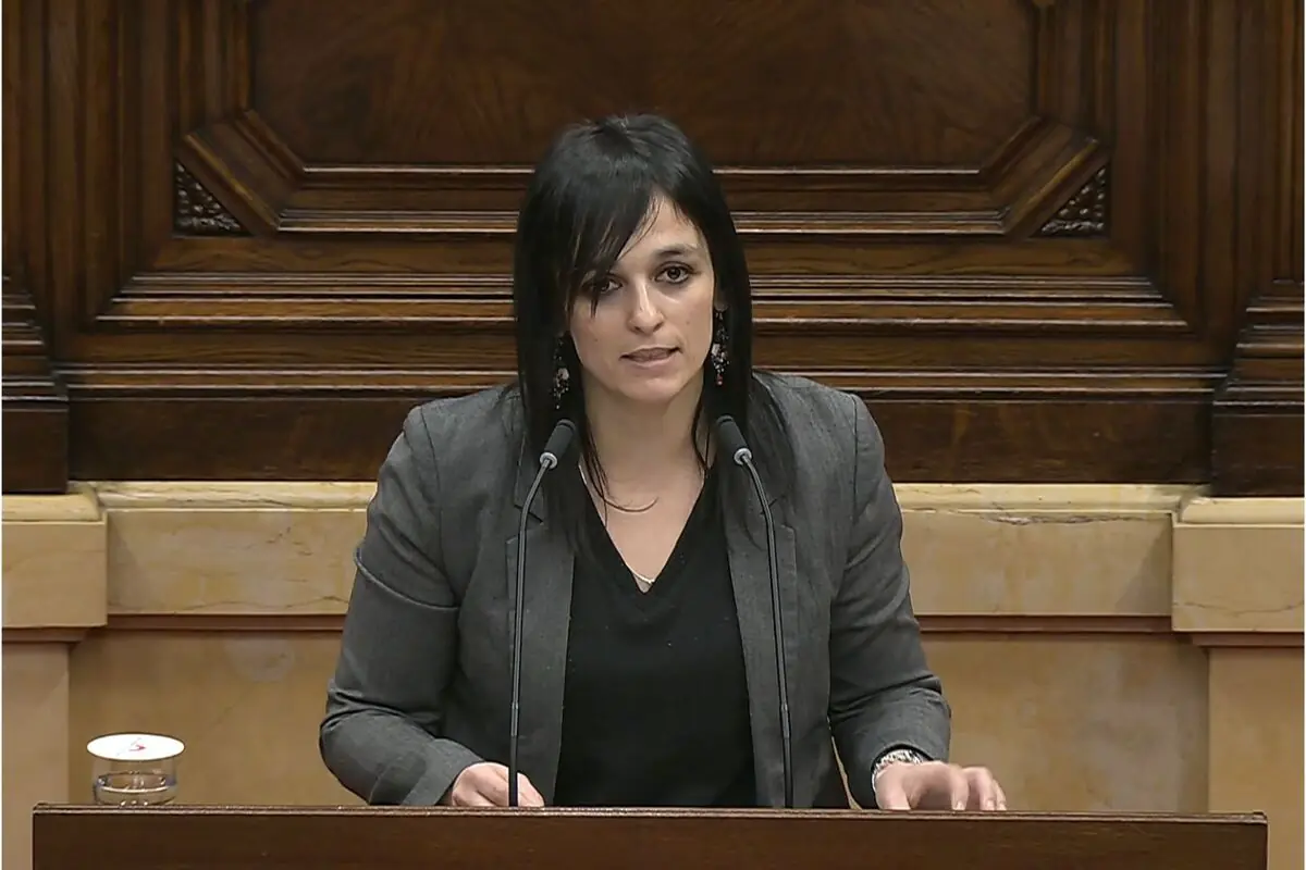 Mujer de cabello oscuro hablando en un atril con dos micrófonos en un salón de plenos de madera