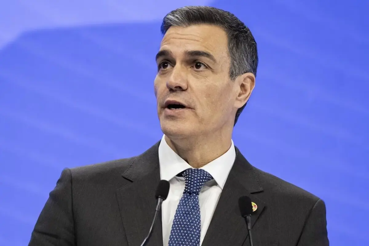 Home amb vestit fosc i corbata blava parlant davant de dos micròfons sobre un fons blau