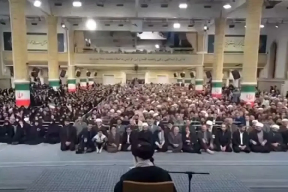 Gran multitud asseguda dins d’un gran saló amb columnes decorades amb els colors de la bandera de l’Iran escoltant un orador situat en primer pla d’esquena a la càmera.