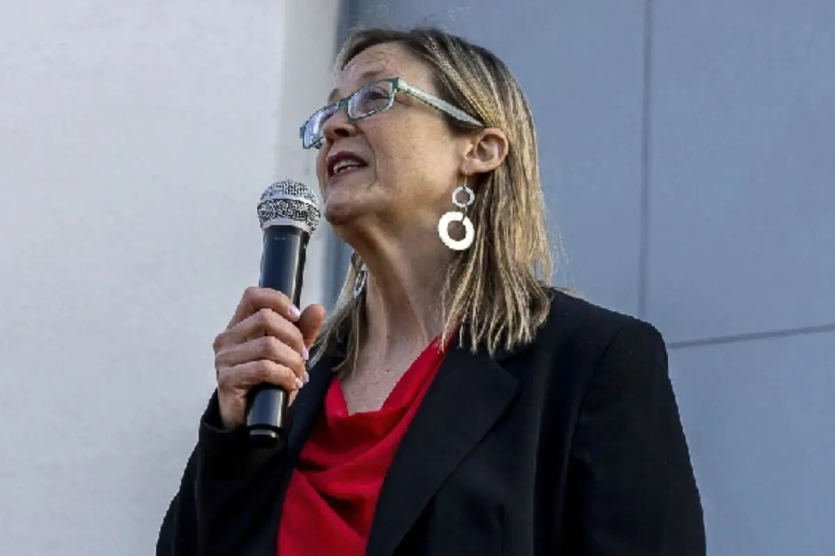 Dona de mitjana edat amb ulleres i arracades circulars parlant per un micròfon mentre porta una jaqueta negra i una brusa vermella