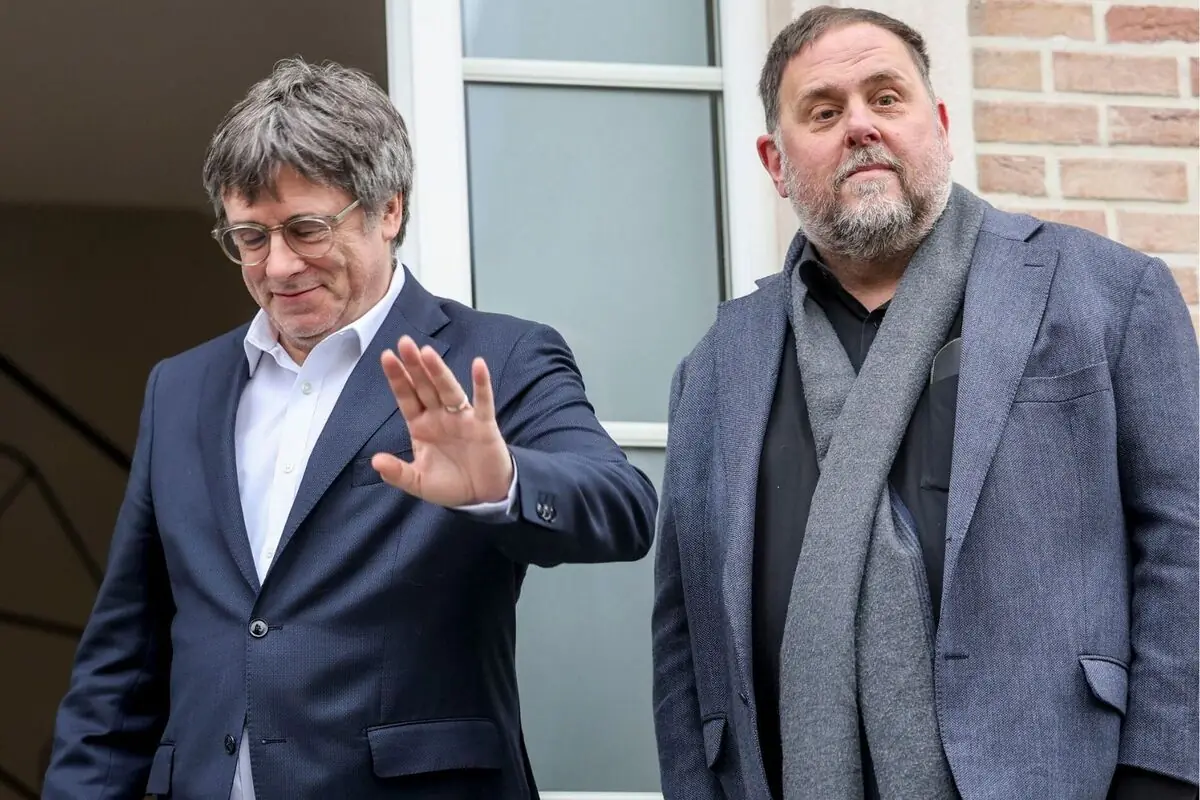 Dos hombres con traje posan juntos frente a un edificio uno de ellos levanta la mano en gesto de saludo