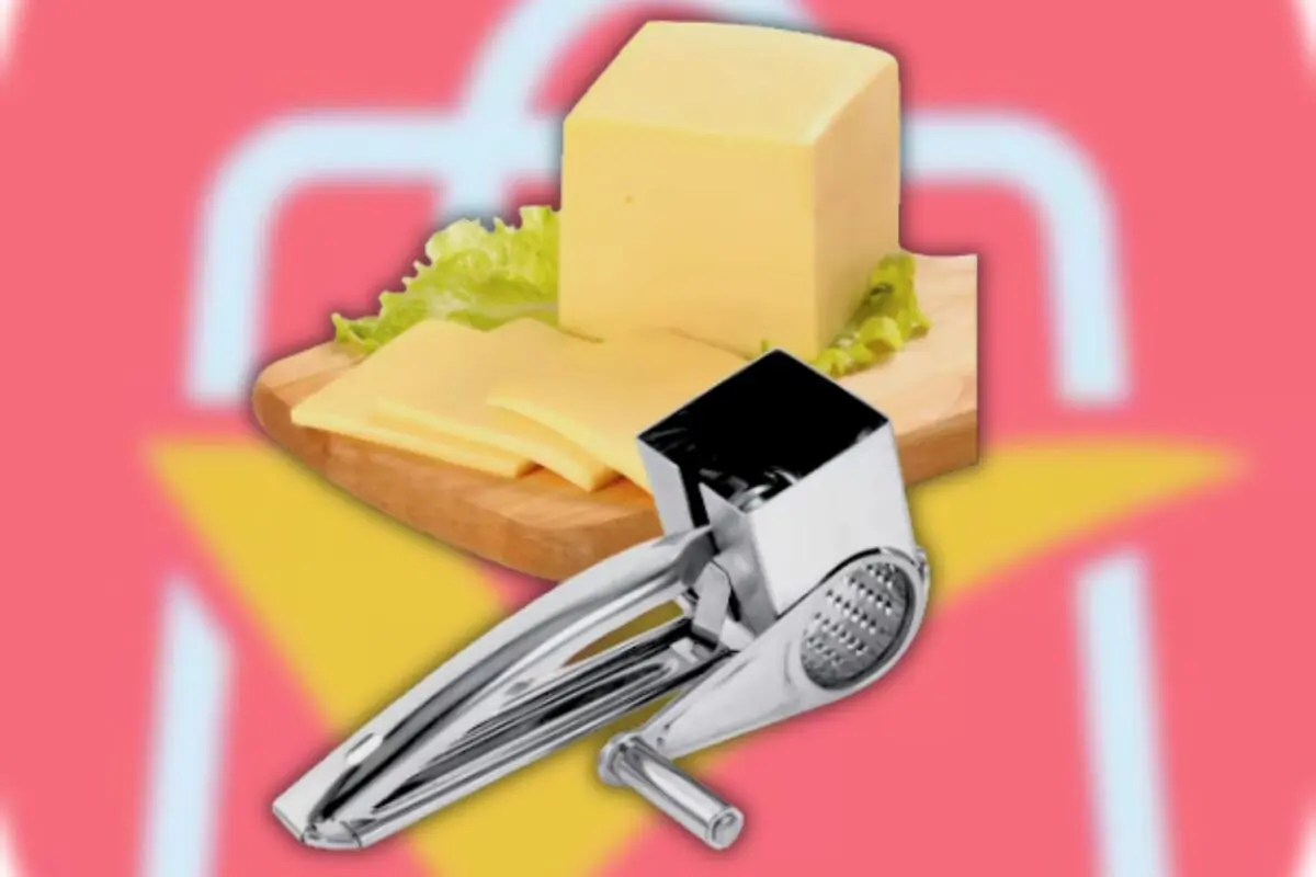 Rallador de queso metálico frente a una tabla de madera con bloque y rebanadas de queso sobre fondo rosa