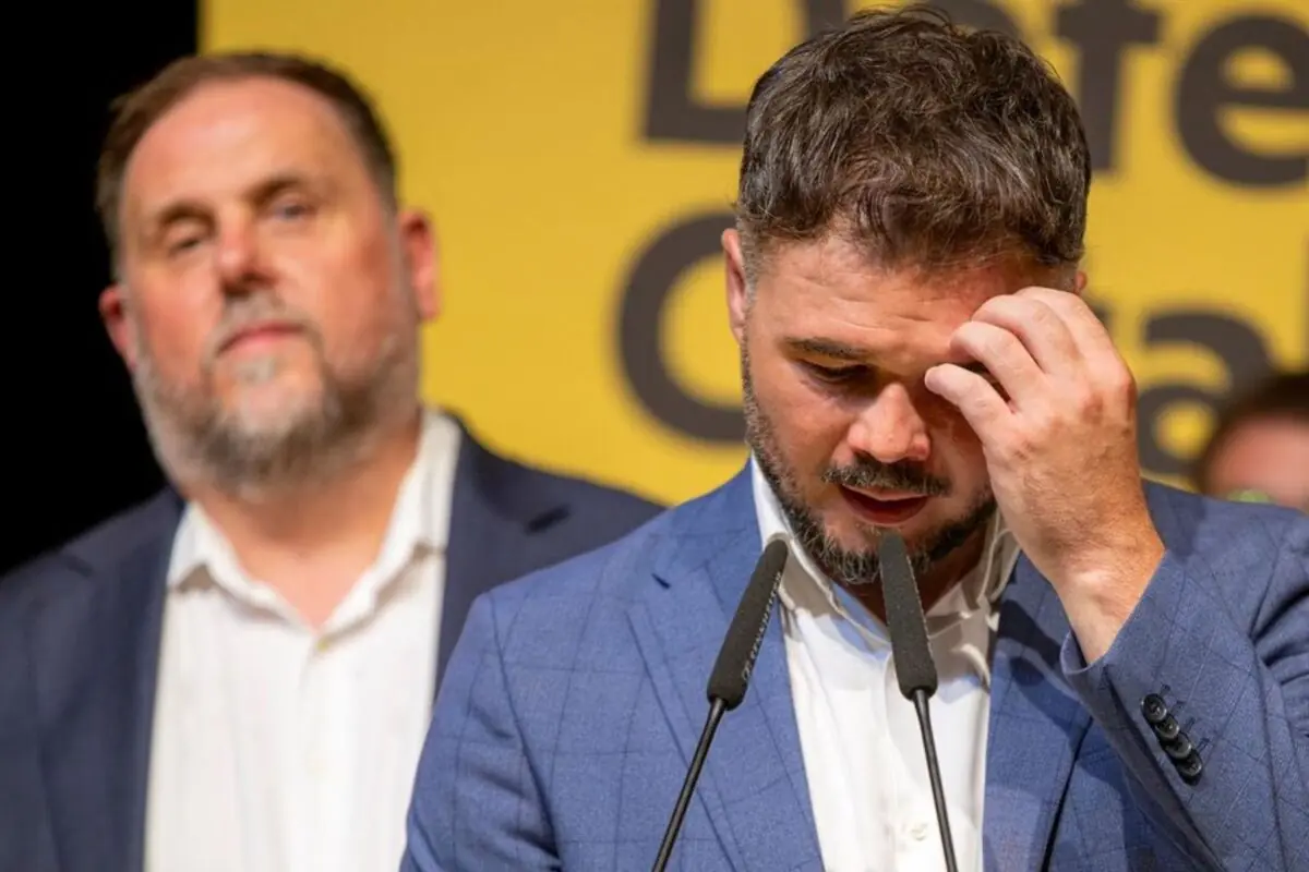 Home amb vestit blau parlant pensatiu davant de dos micròfons mentre una altra persona desenfocada roman al darrere sobre un fons groc amb text incomplet