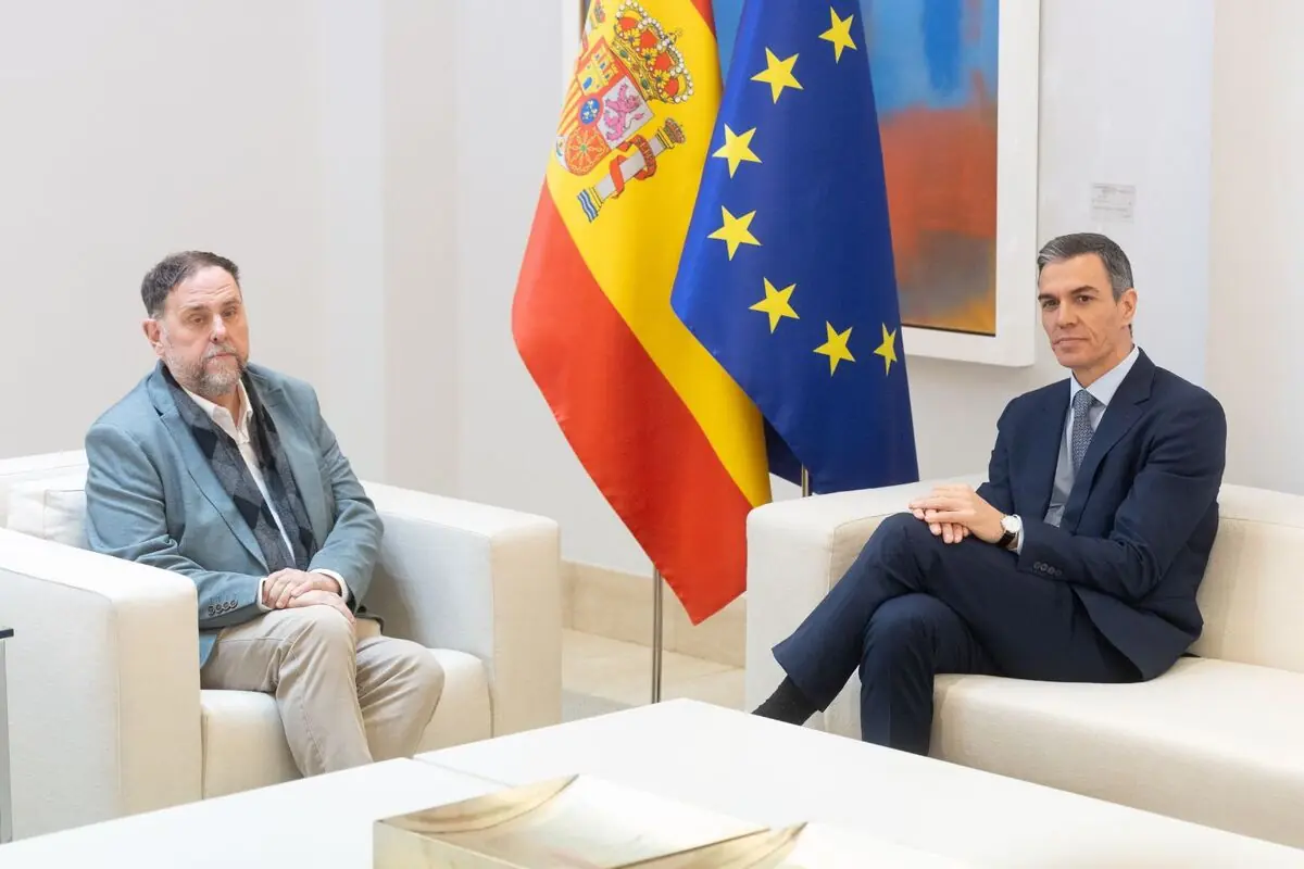 Dos homes amb vestit asseguts en butaques blanques l’un davant de l’altre en una sala de reunions amb les banderes d’Espanya i de la Unió Europea al fons