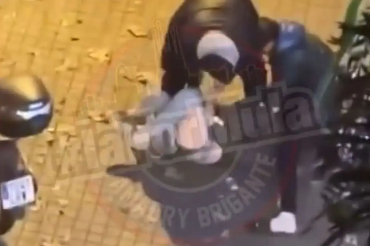 Persona con casco y chaqueta oscura agachada junto a una moto en una acera de noche con hojas secas en el suelo