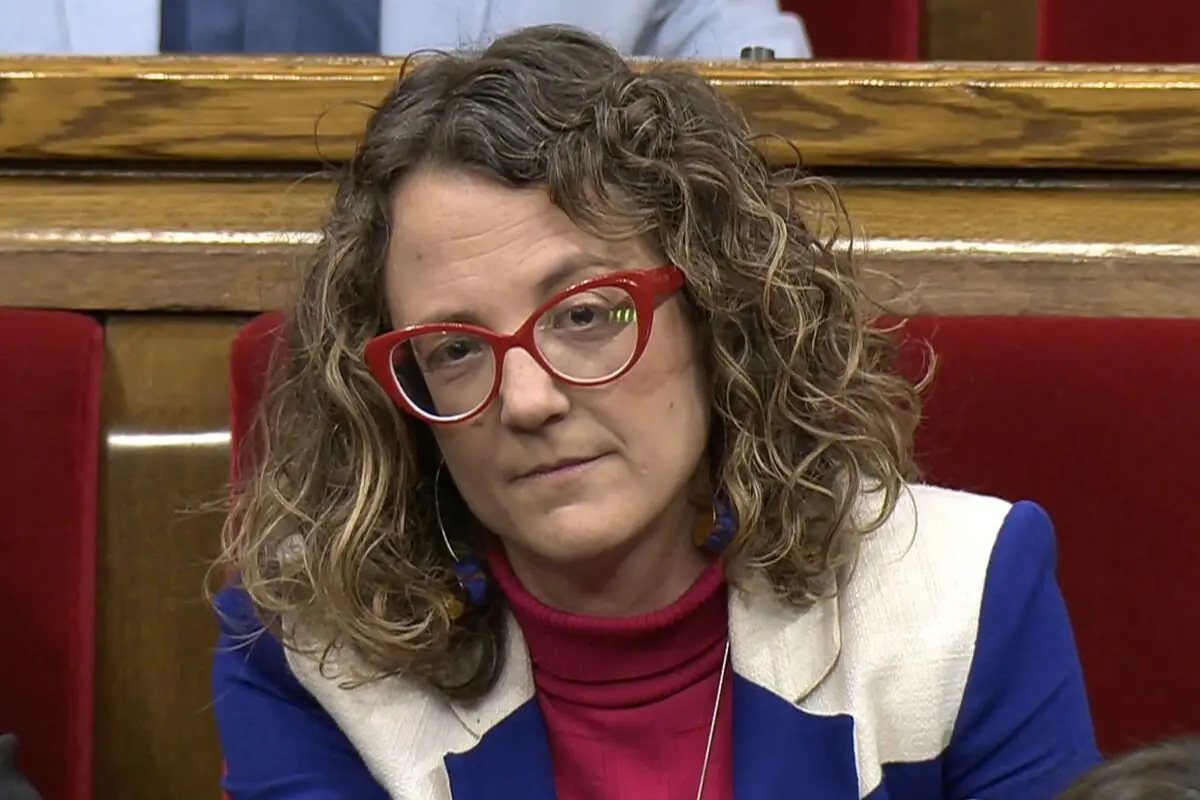 Mujer de cabello rizado con gafas rojas sentada en un asiento rojo de un salón institucional mirando hacia la cámara