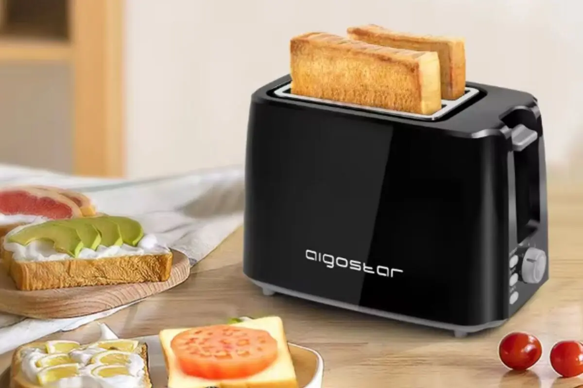 Tostadora eléctrica negra con dos rebanadas de pan tostándose sobre una mesa de desayuno con tostadas y frutas alrededor