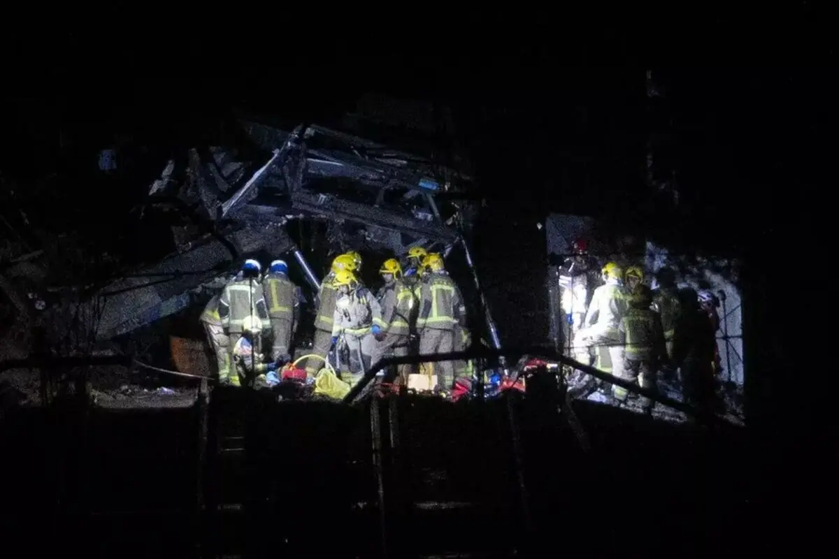 Bomberos con cascos y trajes reflectantes trabajan de noche entre escombros iluminados por focos en una zona de rescate