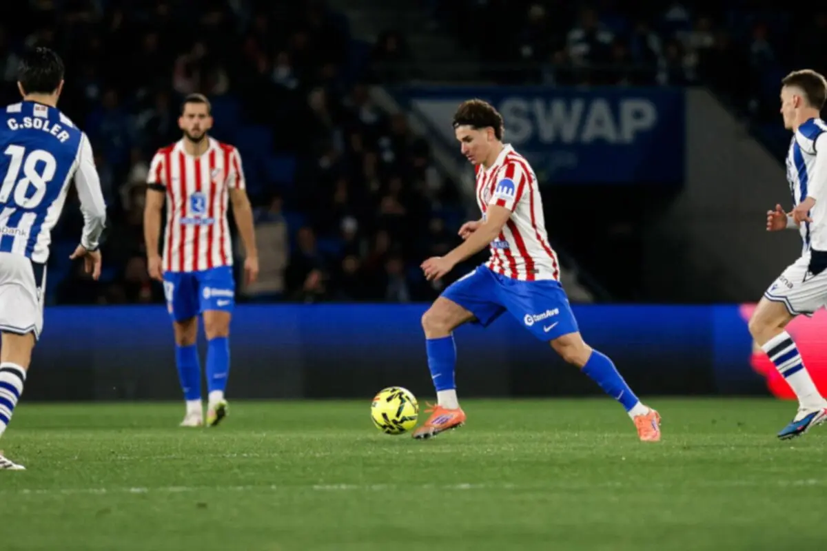 Futbolista amb samarreta blanc-i-vermella i pantaló blau condueix la pilota en un partit mentre rivals amb uniforme blanc i blau s’acosten per pressionar-lo Futbolista amb samarreta blanc-i-vermella i pantaló blau condueix la pilota en un partit mentre rivals amb uniforme blanc i blau s’acosten per pressionar-lo