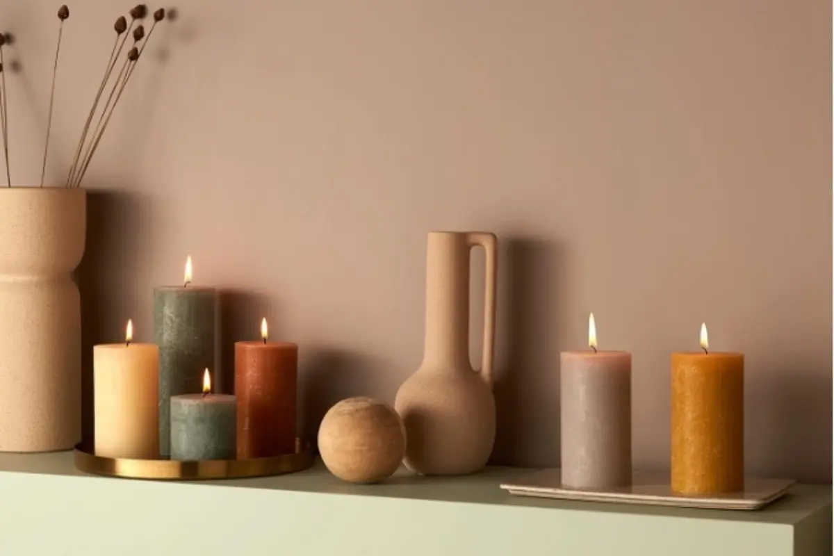 Velas encendidas de colores cálidos sobre una repisa junto a jarrones y una esfera decorativa en un ambiente minimalista