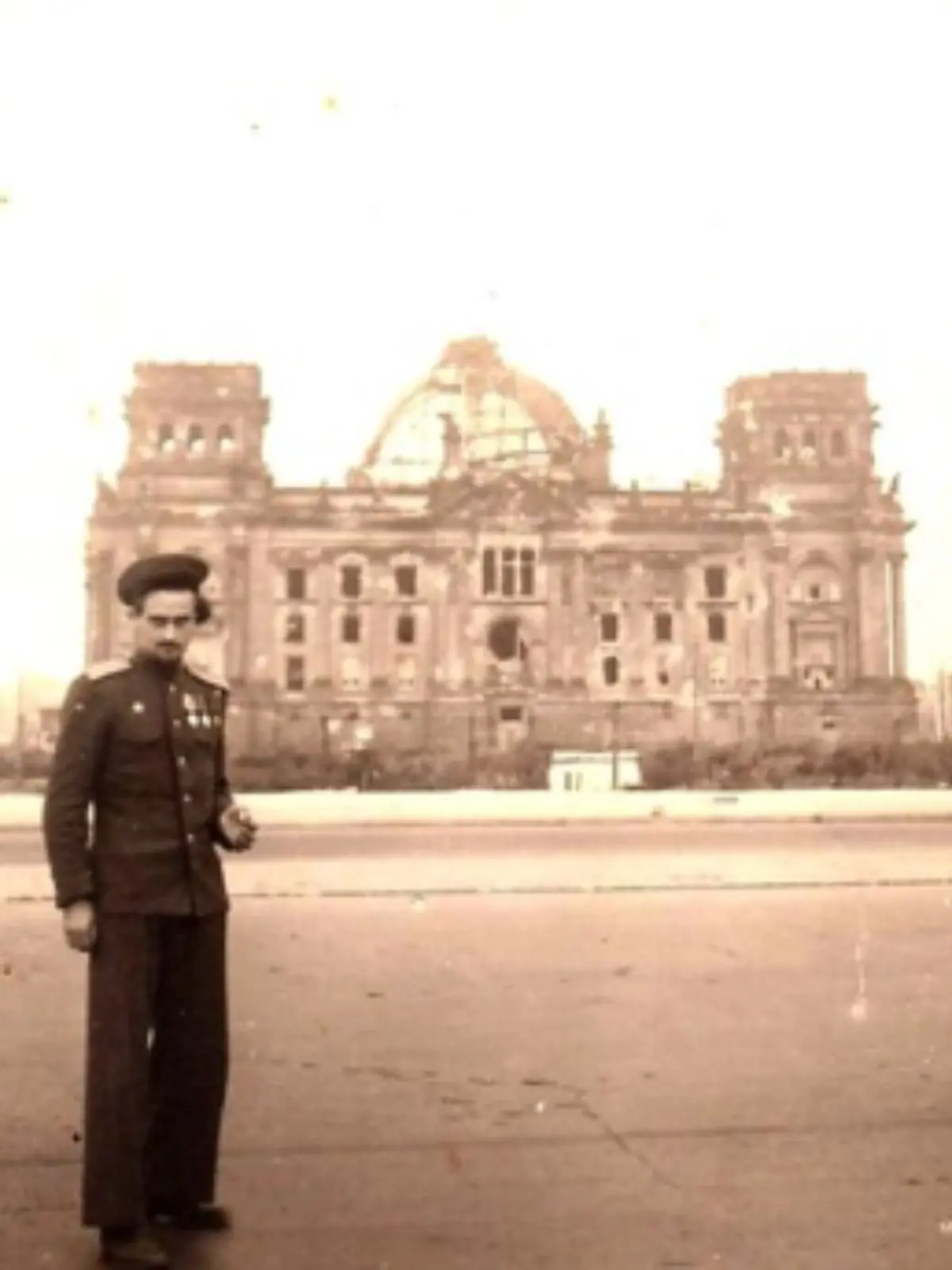 Vladímir Natánovitx Gelfand amb uniforme militar posant en un carrer ample davant d’un gran edifici històric danyat i ennegrit per la guerra.