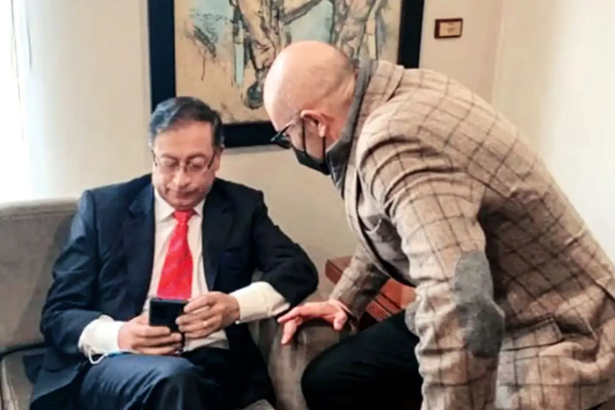 Dos hombres con traje conversan en una sala mientras uno de ellos mira la pantalla de su teléfono móvil Dos hombres con traje conversan en una sala mientras uno de ellos mira la pantalla de su teléfono móvil