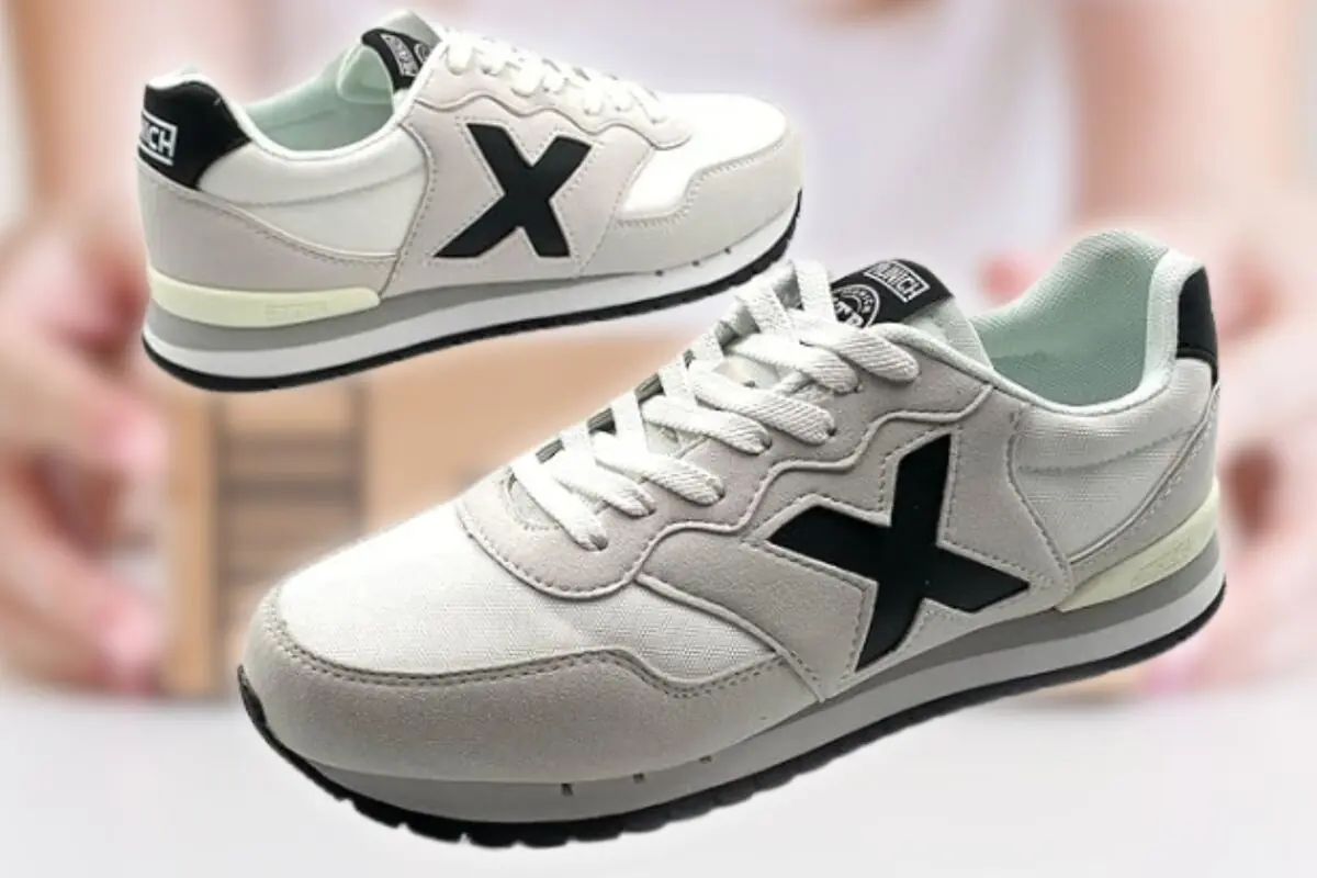 Zapatillas deportivas blancas con suela gris y detalle de gran X negra en el lateral sobre fondo desenfocado