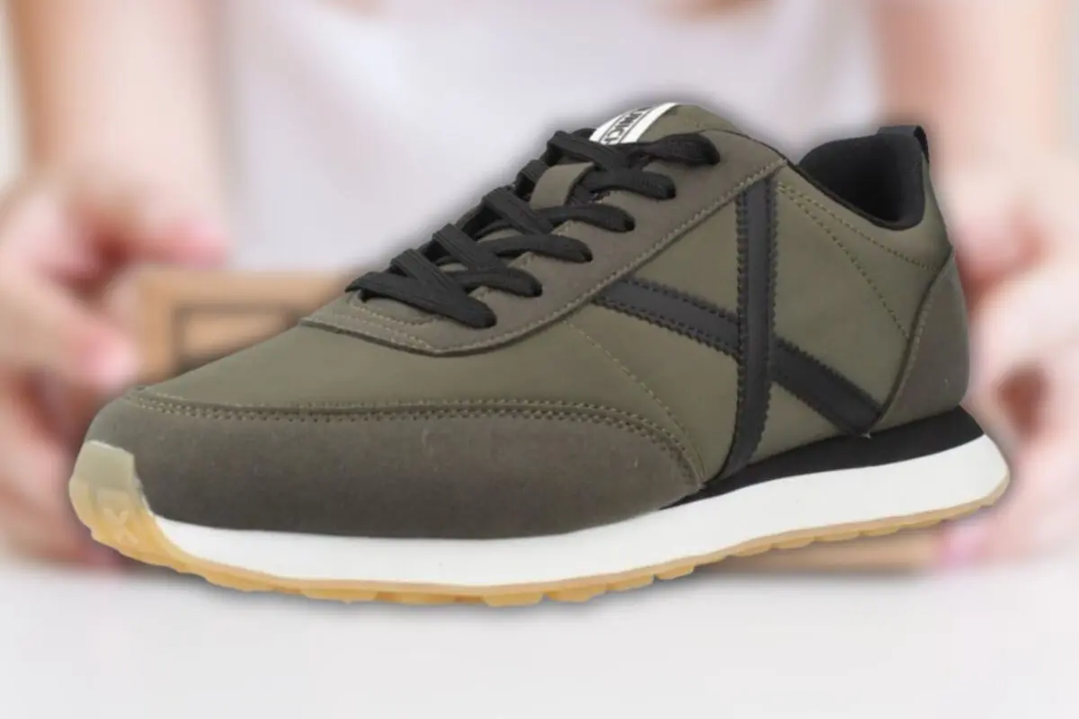 Zapatilla deportiva verde oliva con cordones negros y suela blanca de goma sobre fondo desenfocado