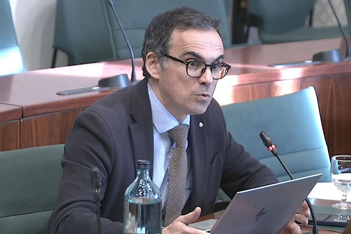 Hombre con gafas y traje hablando frente a un micrófono mientras usa una computadora portátil en una sala de reuniones