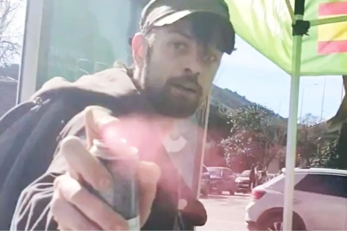 Hombre con gorra y barba usando un aerosol hacia la cámara en una calle con autos y carpa verde de fondo