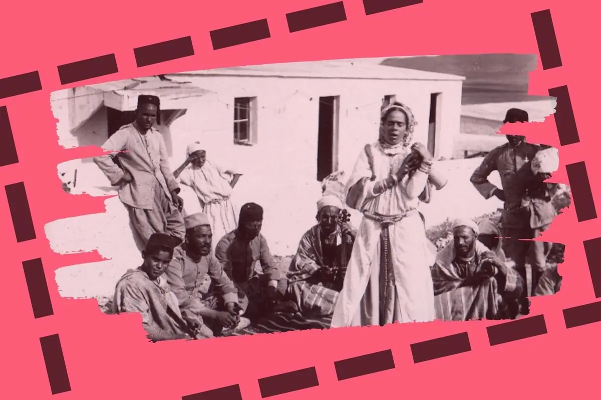 Grup de persones amb vestimenta tradicional reunides a l’aire lliure mentre una dona dreta sembla cantar o parlar en primer pla sobre un fons rosa amb un marc decoratiu.