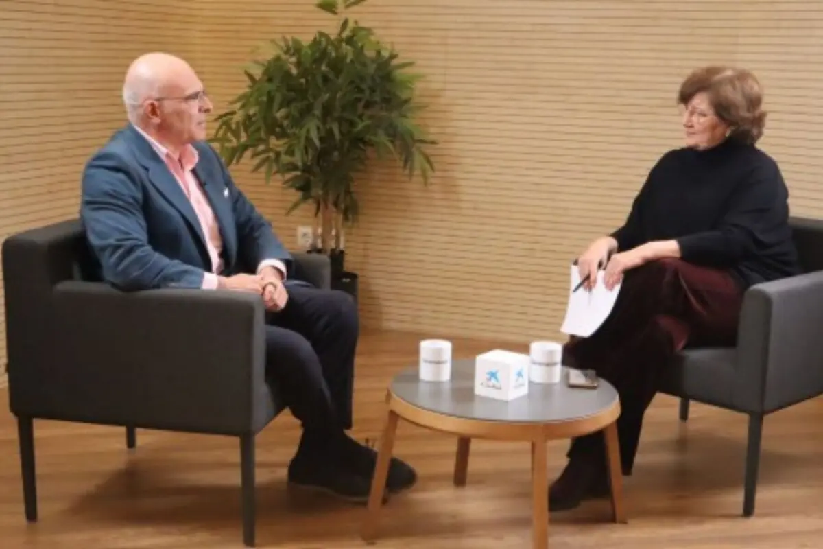 Dues persones assegudes en butaques grises conversen en una sala amb una taula baixa al centre i una planta decorativa al fons