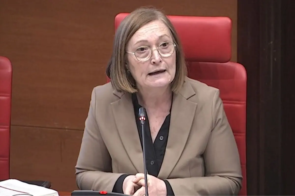 Dona de mitjana edat amb ulleres i americana beix parlant davant d’un micròfon en una sala de conferències amb seients vermells