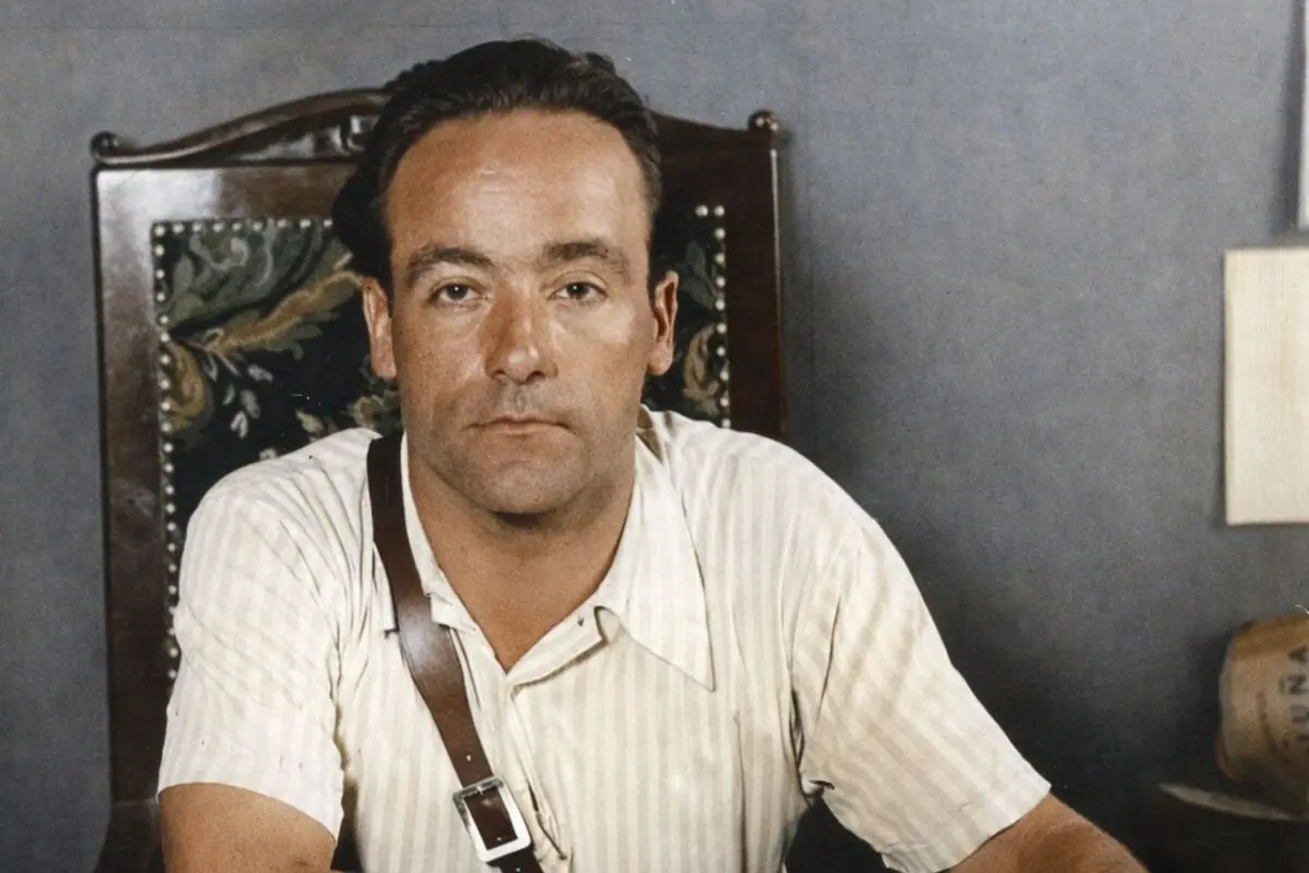 Joan García Oliver amb camisa clara i tirant de cuir assegut en una cadira de fusta tallada mirant a la càmera.