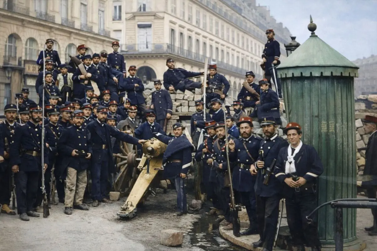 Grup nombrós de soldats amb uniformes blaus i boines vermelles posant al costat d’un canó i una barricada de pedres en un carrer d’una antiga ciutat europea.