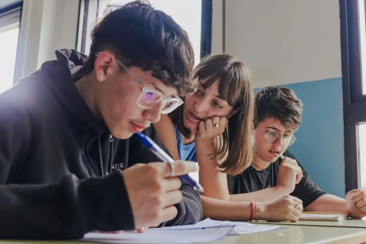 Tres estudiantes adolescentes con gafas trabajando concentrados en sus cuadernos en un aula iluminada por la ventana
