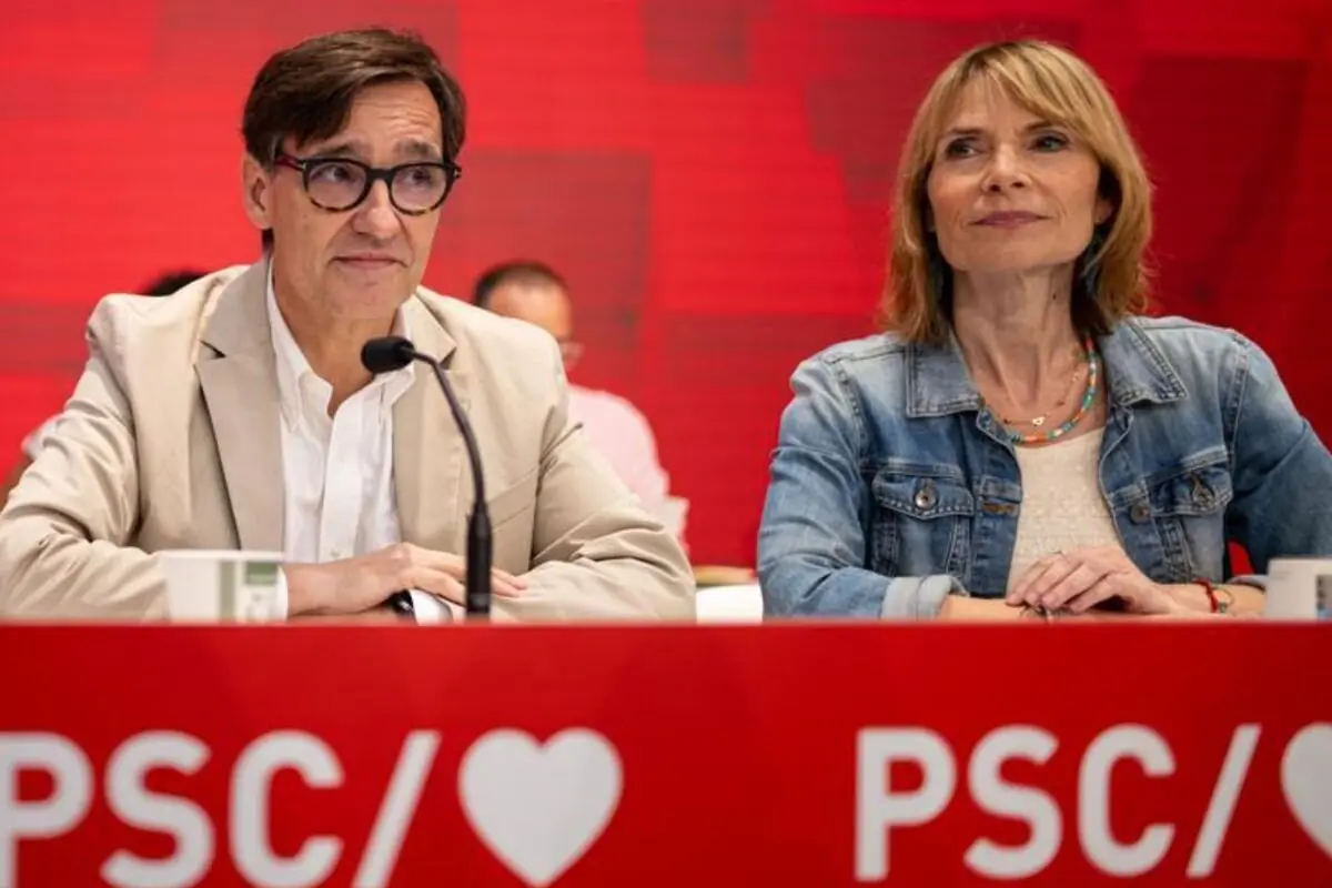 Dues persones assegudes darrere d’una taula vermella amb el logotip PSC i un cor en un acte o roda de premsa davant d’un fons vermell Dues persones assegudes darrere d’una taula vermella amb el logotip PSC i un cor en un acte o roda de premsa davant d’un fons vermell