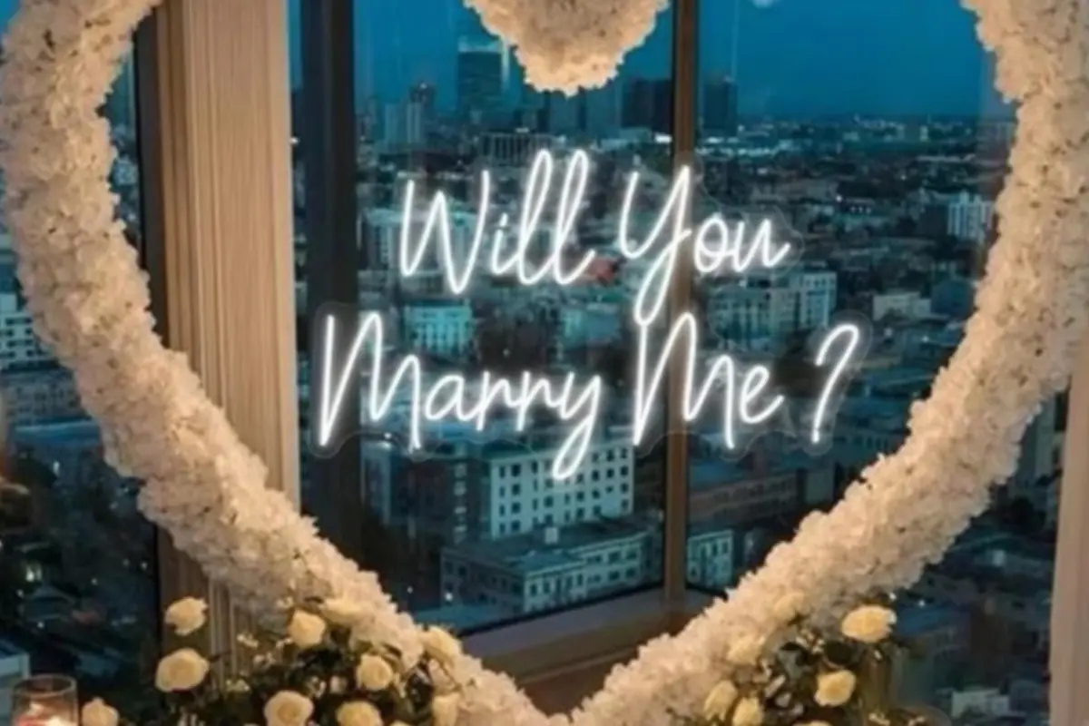 Escena romántica de propuesta de matrimonio con un gran corazón de flores blancas frente a una ventana con vista nocturna a la ciudad y un letrero de neón que dice 'Will you marry me?'