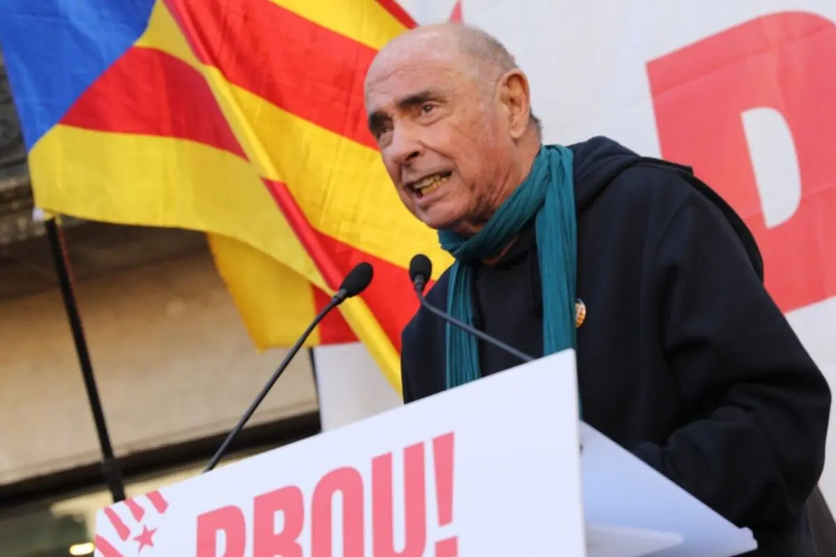 Hombre mayor hablando en un mitin político al aire libre con micrófonos y una bandera catalana de fondo