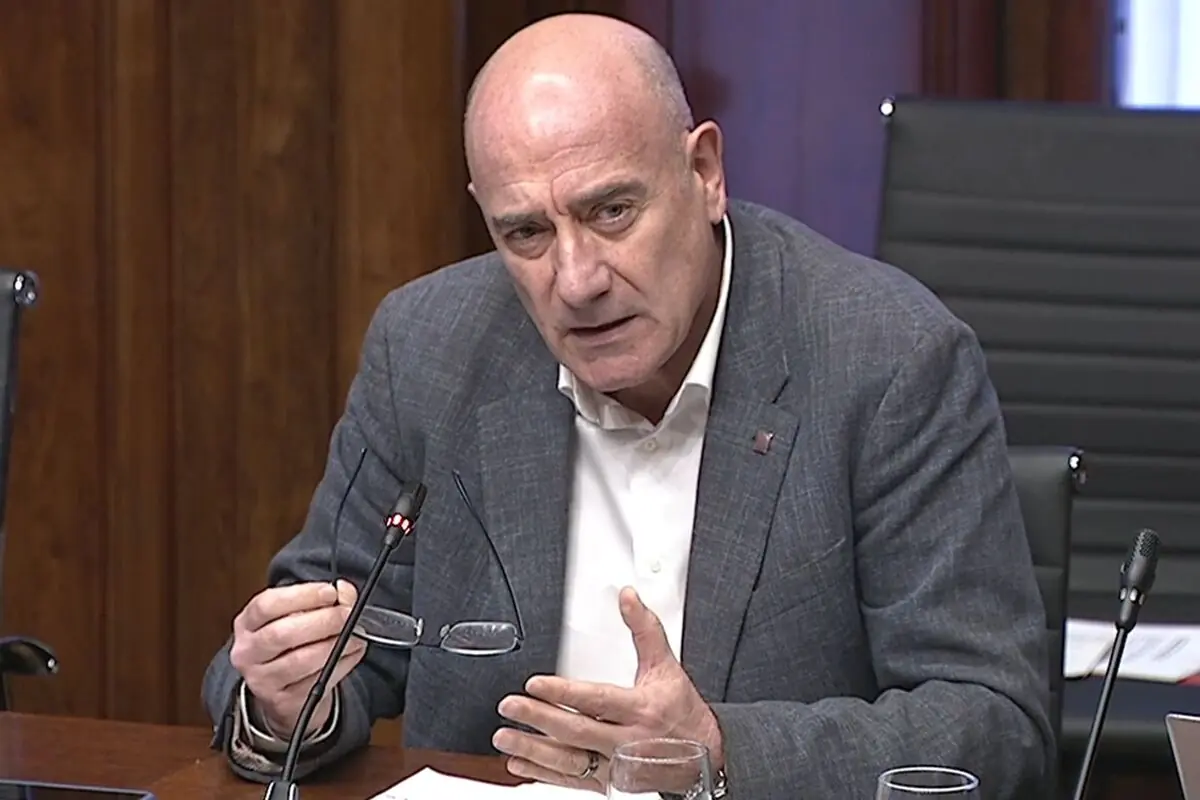 Home calb de mitjana edat amb vestit gris parlant en una sala de reunions mentre sosté unes ulleres i un micròfon davant seu