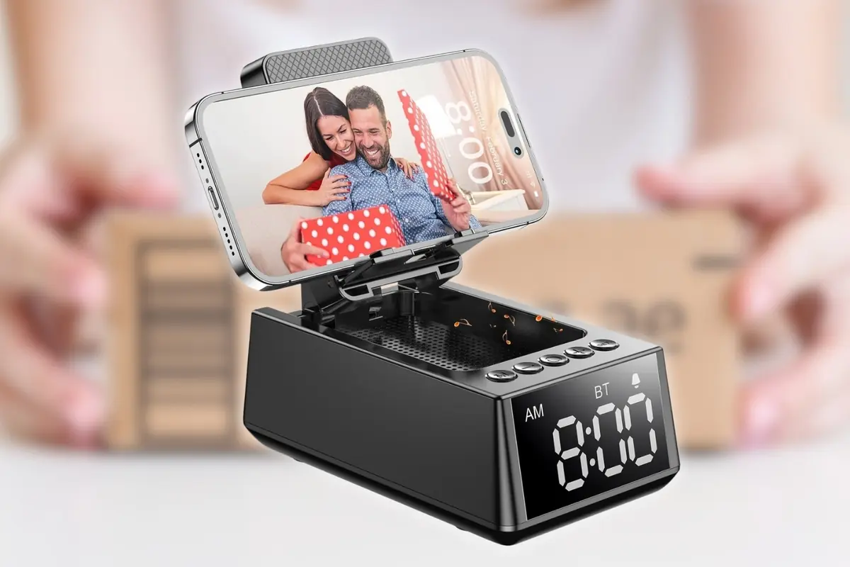 Reloj despertador digital negro con base multifunción y altavoz que sostiene un smartphone en posición horizontal mostrando a una pareja feliz abriendo un regalo
