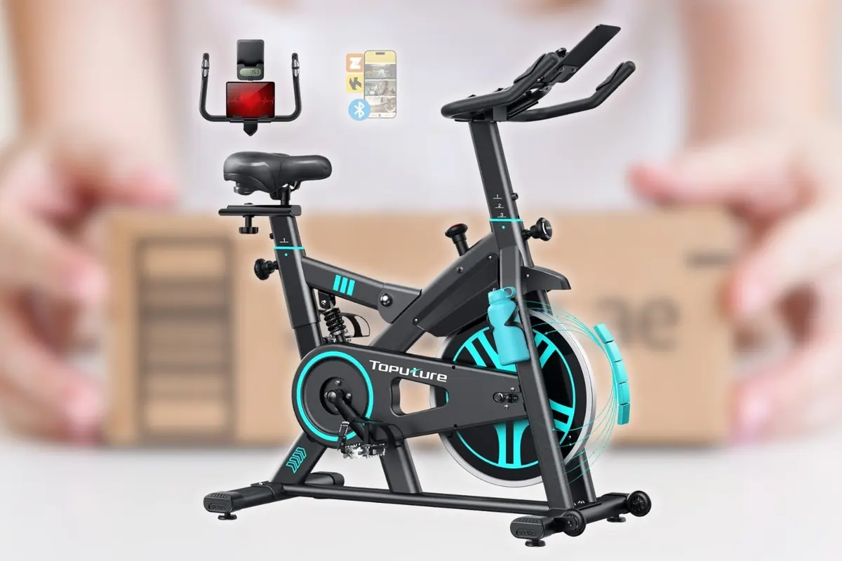 Bicicleta estática de interior negra con detalles en azul turquesa marca Toputure con soporte para tablet y botella frente a un fondo desenfocado de paquete de envío