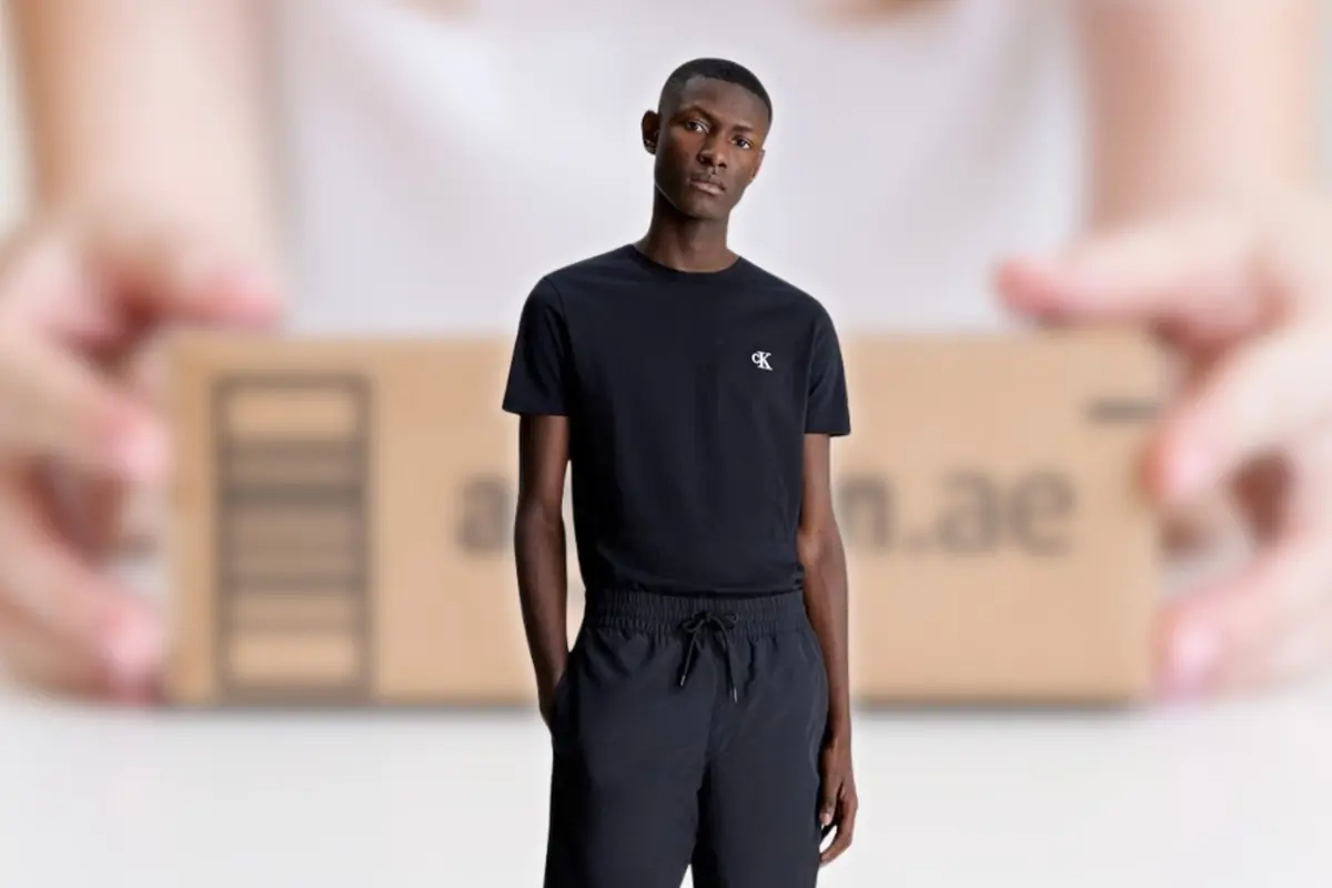 Hombre joven con camiseta y pantalón negros posando frente a una caja de cartón de envío desenfocada en segundo plano