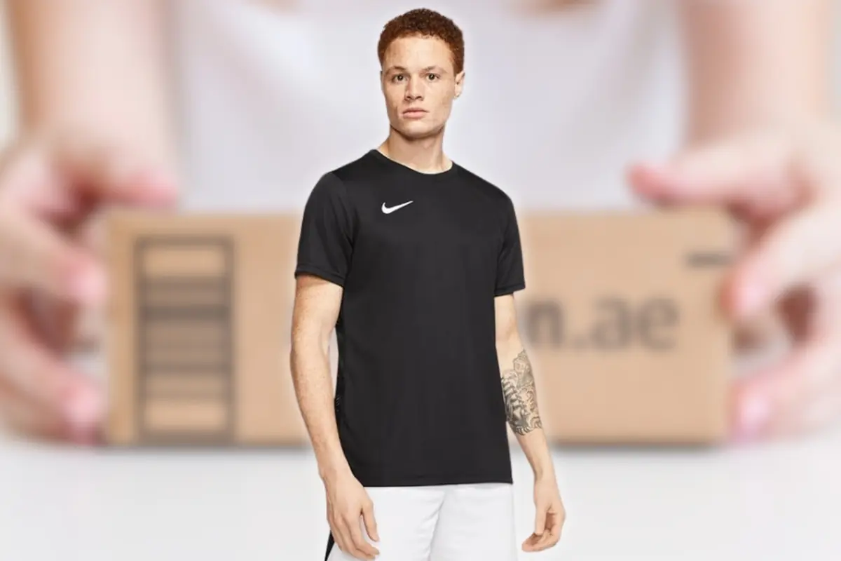 Hombre joven con camiseta deportiva negra y pantalones cortos blancos posando frente a una caja de cartón desenfocada sostenida por unas manos