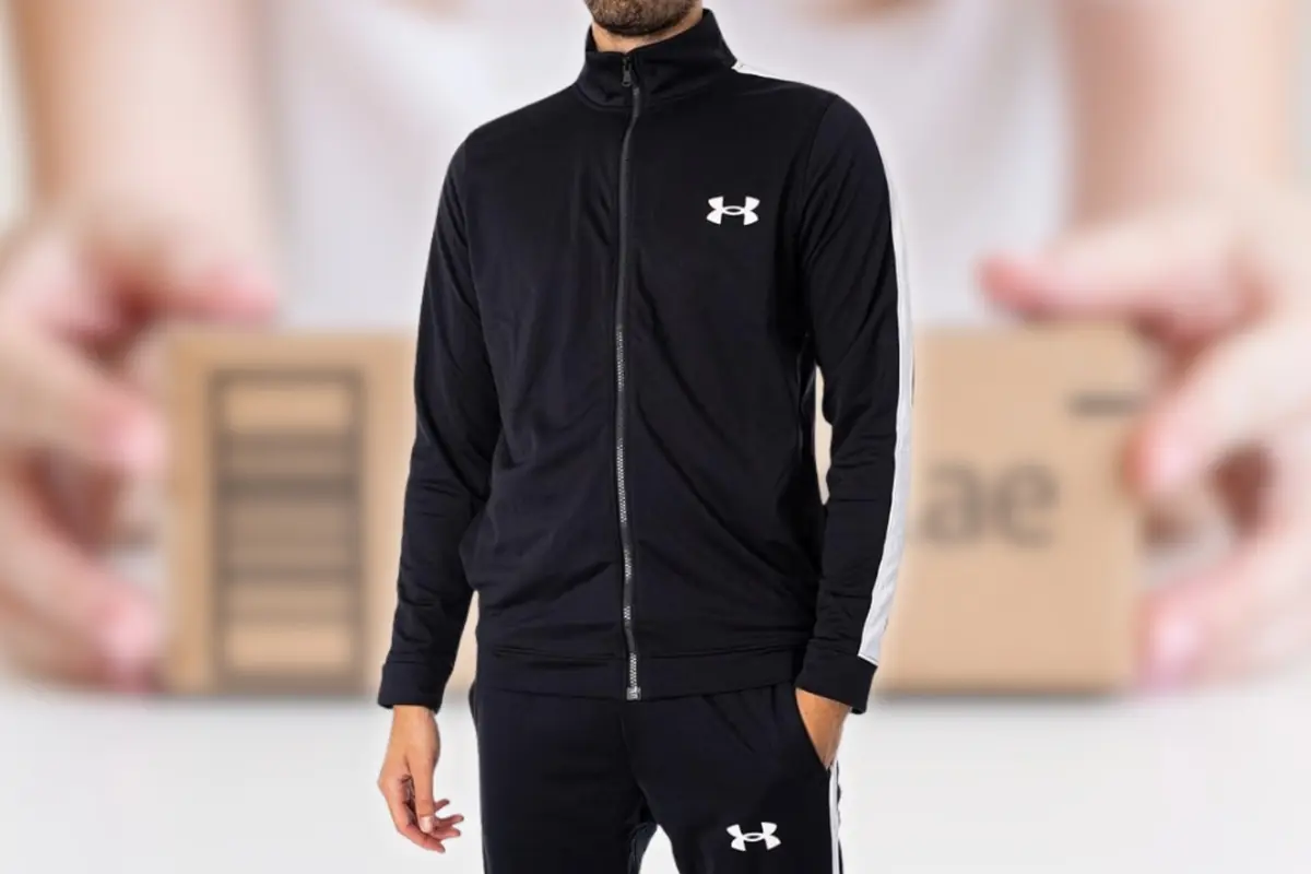 Hombre vistiendo chándal negro deportivo con cremallera y franjas blancas en las mangas sobre fondo desenfocado con caja de cartón