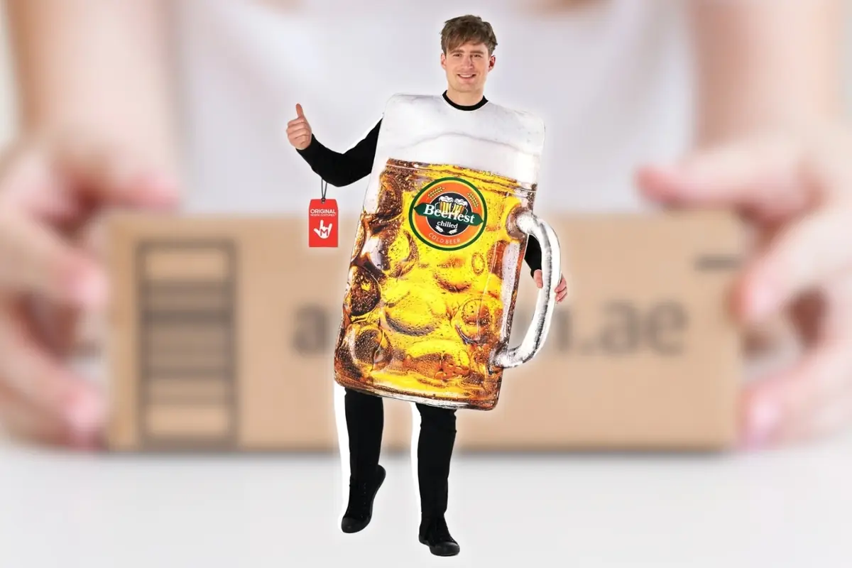 Montaje con hombre disfrazado de jarra de cerveza gigante con espuma y asa plateada posando sonriente frente a una caja de cartón desenfocada