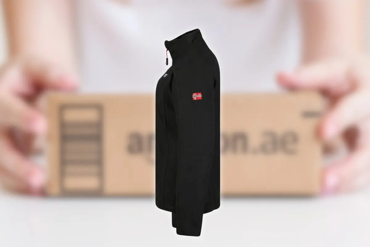 Vista lateral de un polar negro con cremallera y pequeño parche de bandera en la manga frente a una caja de envío de comercio electrónico desenfocada en el fondo