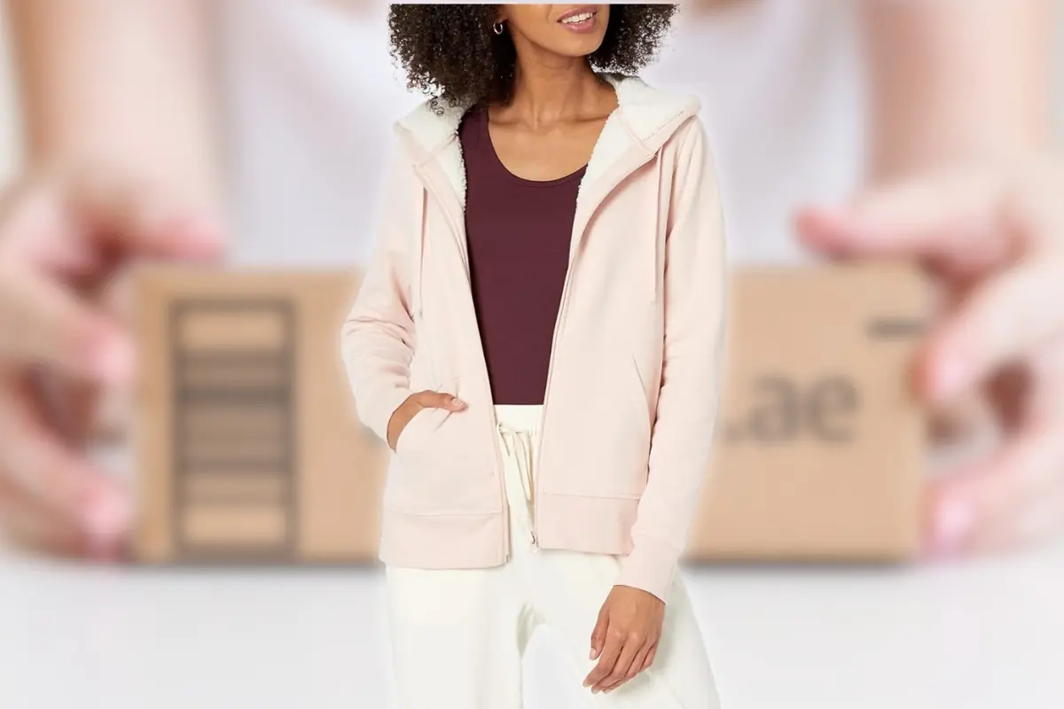 Mujer con sudadera rosa con capucha y forro de borreguito sobre camiseta burdeos y pantalón claro con cajas de envío desenfocadas al fondo