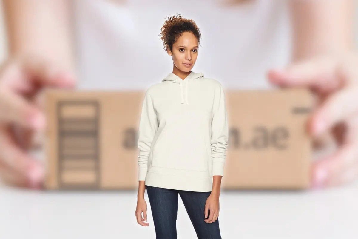 Mujer joven con sudadera beige y vaqueros de pie frente a una caja de cartón desenfocada sostenida por unas manos